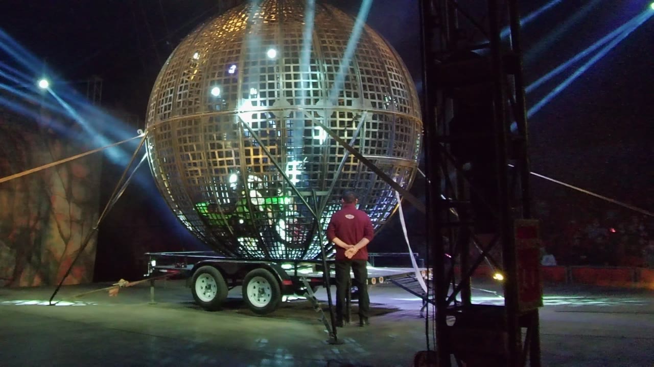 At 0:00 in San Jose Del Cabo, a large reflective grid sphere sits on a trailer while a person in a red shirt stands near the right side under blue stage lights.