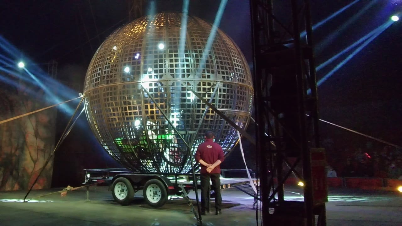 At 0:02 in San Jose Del Cabo, the metallic grid sphere fills much of the frame, with the person still beside it and blue spotlights projecting from the dark stage area.