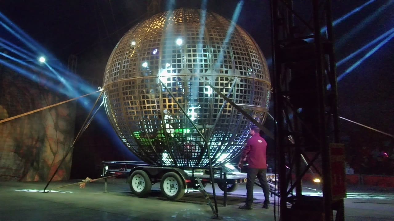 In San Jose del Cabo, a large golden disco ball on a wheeled trailer fills the stage area while a person in a red shirt stands near its support frame under blue lights.