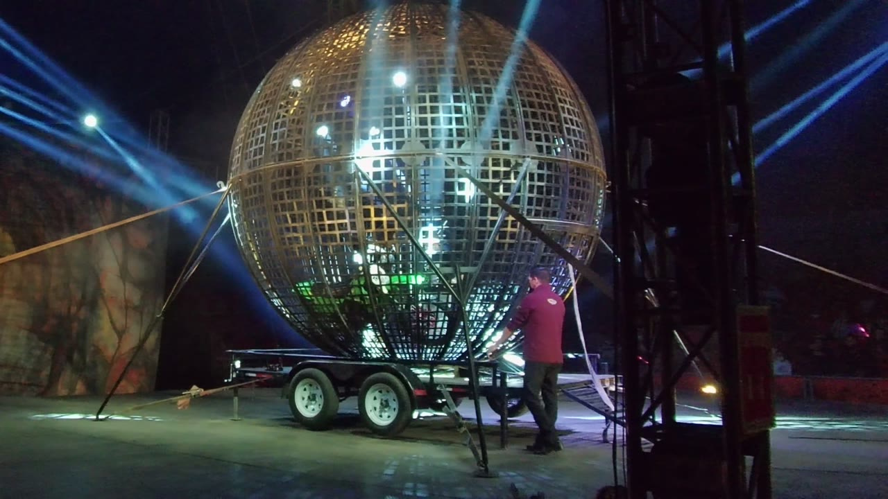 Person in a red shirt adjusts equipment beside a large disco ball on a trailer.