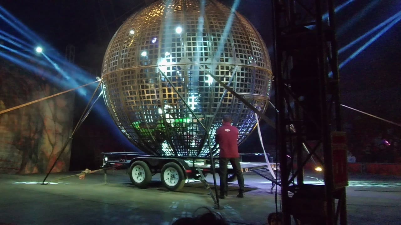In San Jose del Cabo, the worker stands at the right side of the golden disco ball trailer while blue stage beams cross behind the rig and dark truss edges frame the view.