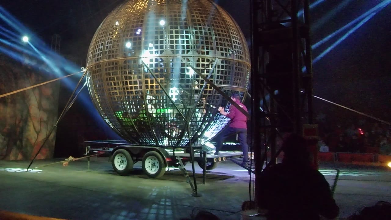 Large golden metal-grid spherical stage prop on a wheeled trailer in San Jose del Cabo, with blue light beams crossing the night stage and no visible people clearly in frame.