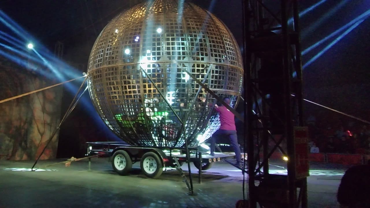 Person in a pink shirt works beside the large golden spherical stage prop on its trailer in San Jose del Cabo, with black truss and blue beams around the stage.