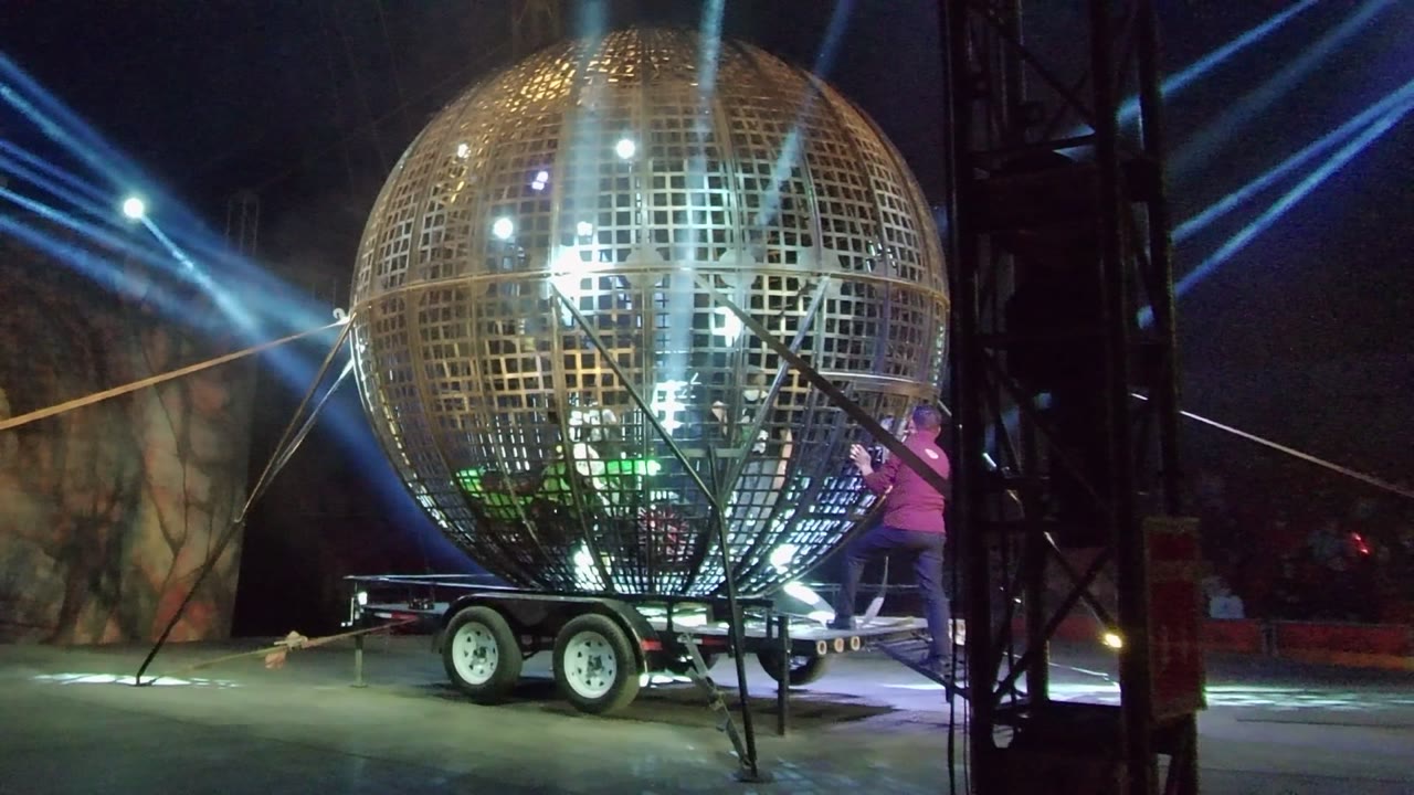 The golden metal-grid sphere dominates the San Jose del Cabo stage view, with trailer wheels below, blue light beams behind, and a person partly visible at the right edge.