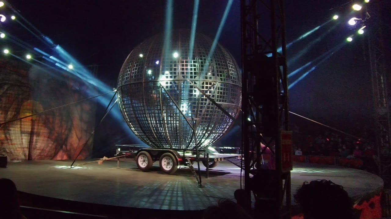 A large grid-pattern spherical stage prop on a trailer is lit by blue and white beams during a San Jose del Cabo night performance, with performers faintly visible inside.