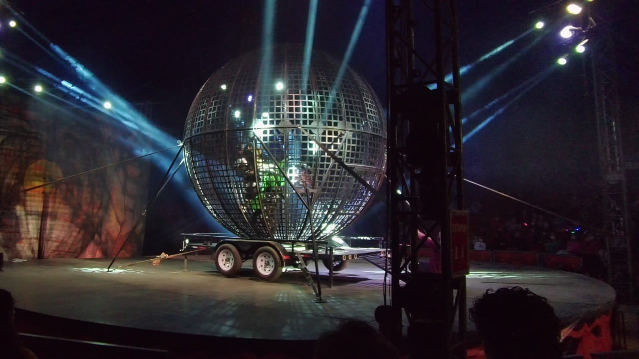 A giant illuminated sphere prop sits slightly left of center on the stage in San Jose del Cabo, with blue beams overhead and dim red points in the dark audience area.