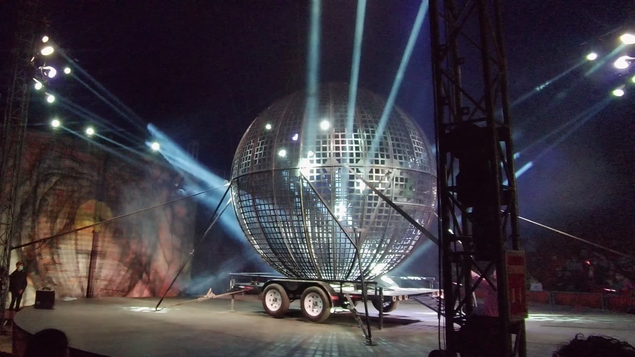 Large gridded metallic sphere on a wheeled stage trailer in San Jose del Cabo, with blue beams crossing the dark stage and a small distant figure near the left edge.