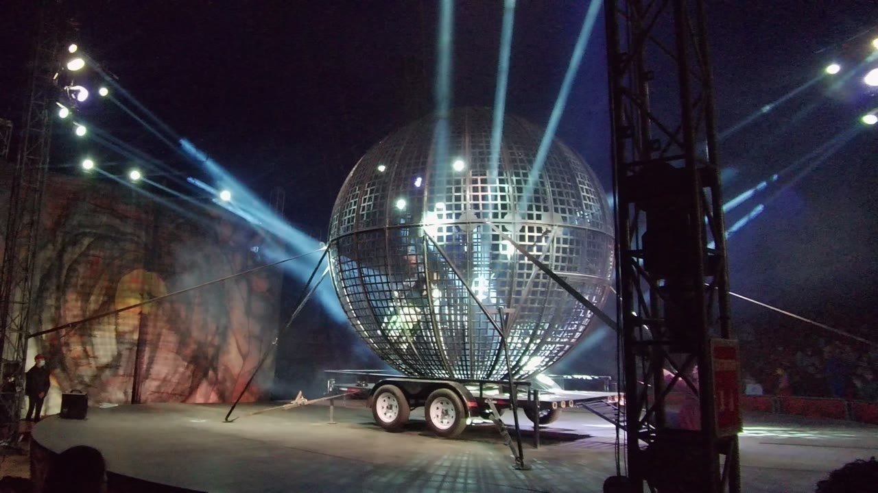 Metallic stage sphere in San Jose del Cabo reflecting bright white overhead lights while blue beams pass behind it and a dark textured backdrop fills the left side.