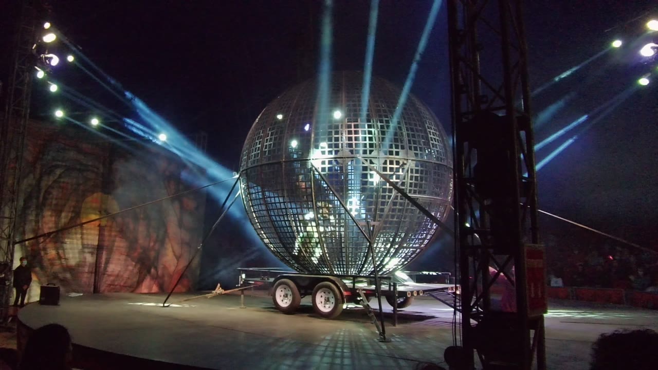 A giant reflective metal sphere on a wheeled trailer sits on a dark stage in San Jose Del Cabo, Mexico, with blue beams overhead and small people visible near the stage edge.