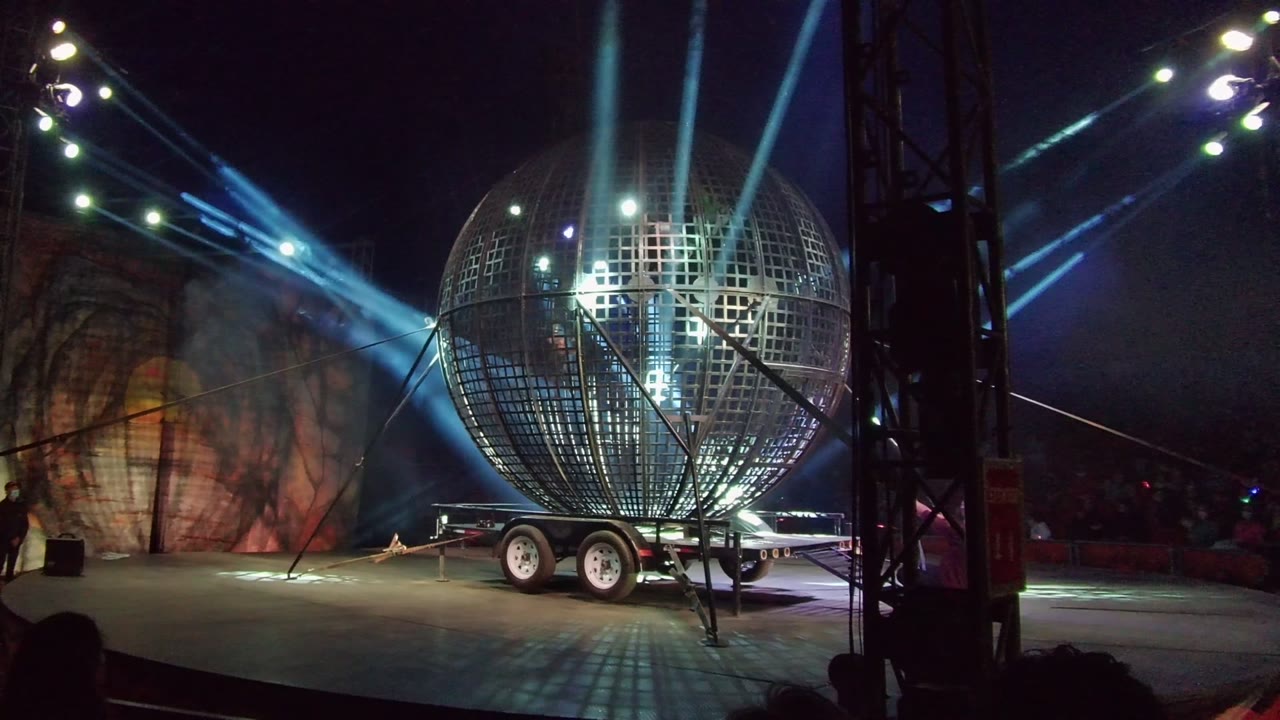 A large mirrored stage sphere on a trailer fills the right side of the frame in San Jose Del Cabo, Mexico, with bright overhead fixtures, blue beams, rigging, and visible people in the dark background.