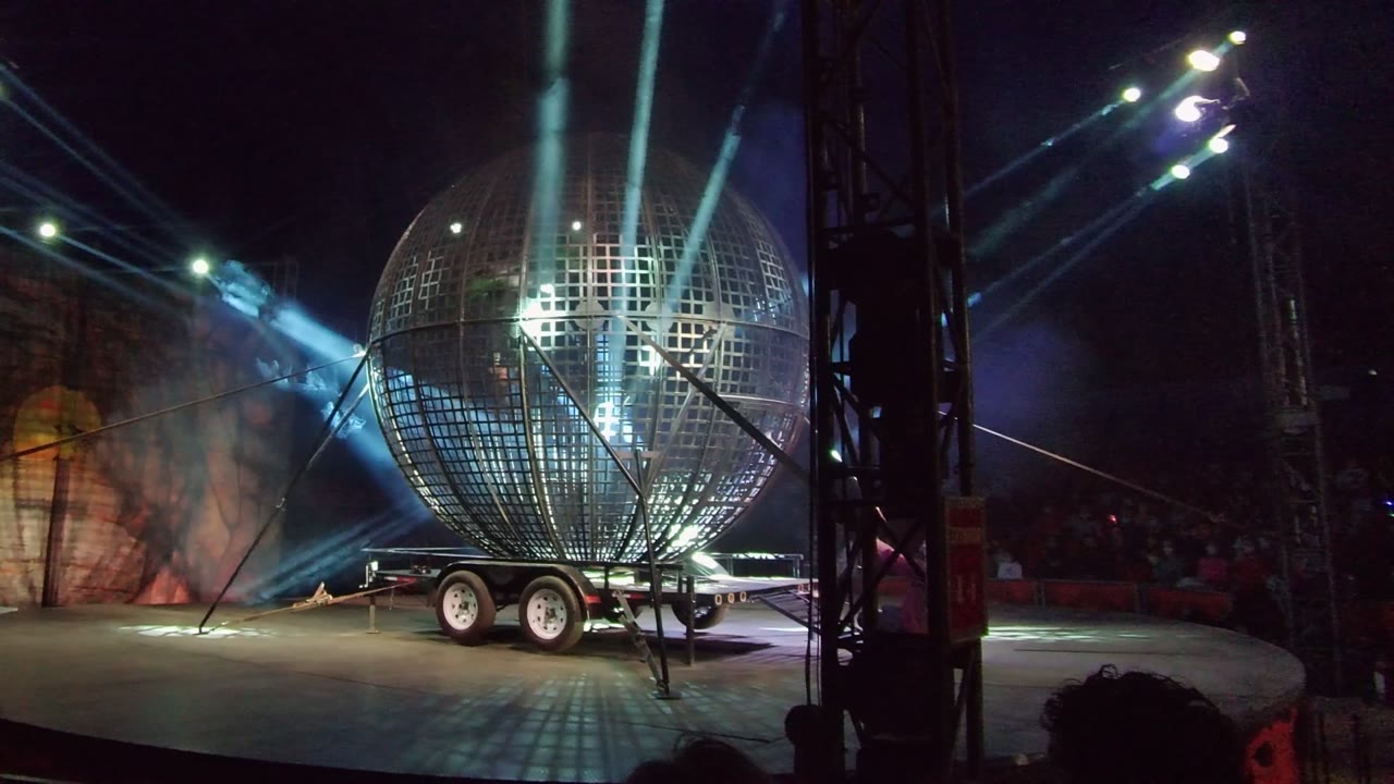 Wide San Jose del Cabo stage view with a large reflective sphere on a trailer, angled rigging cables, blue-white light beams, haze, and audience silhouettes along the lower edge.