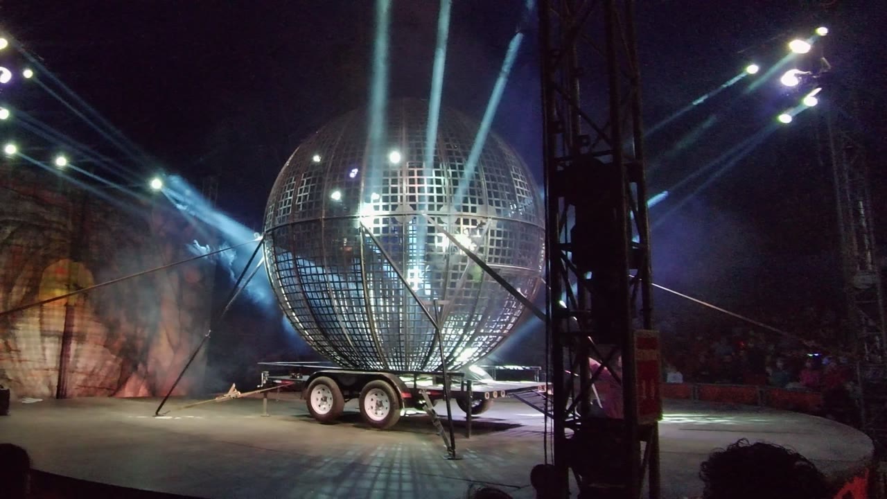 Wide San Jose del Cabo performance stage with the reflective sphere shifted toward the right side of frame, cool light beams in the haze, warm scenic wall panels, and audience silhouettes below.