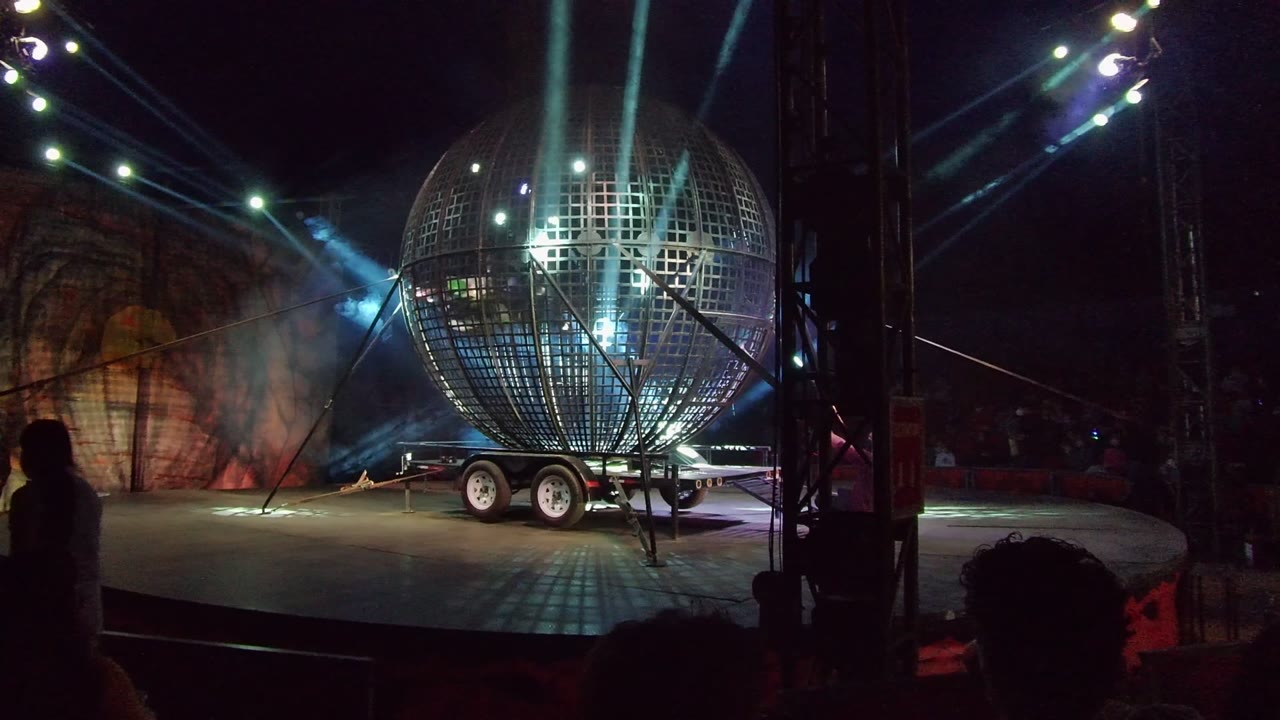 Giant spherical stage prop on a wheeled trailer in San Jose Del Cabo, Mexico, lit by blue-white beams with audience silhouettes along the bottom edge.