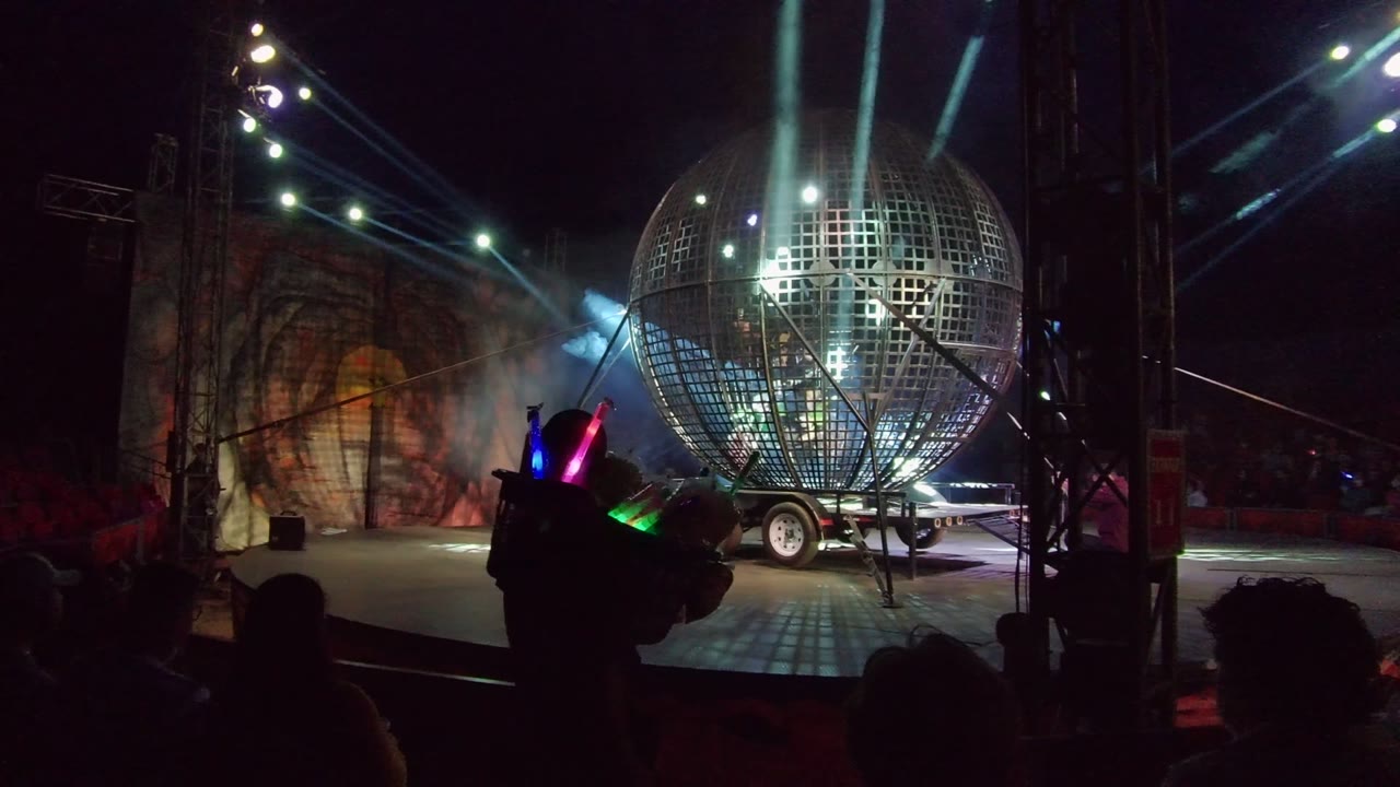 A San Jose Del Cabo stage shows a giant mirrored disco ball on a trailer under blue-white beams, with audience silhouettes and glowing sticks in the foreground.