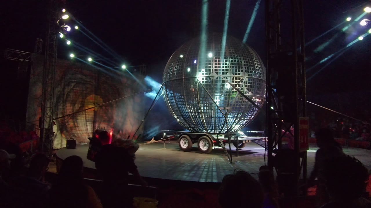 A giant disco ball on its trailer dominates the San Jose Del Cabo stage as overhead spotlights send bright beams through haze above audience silhouettes.