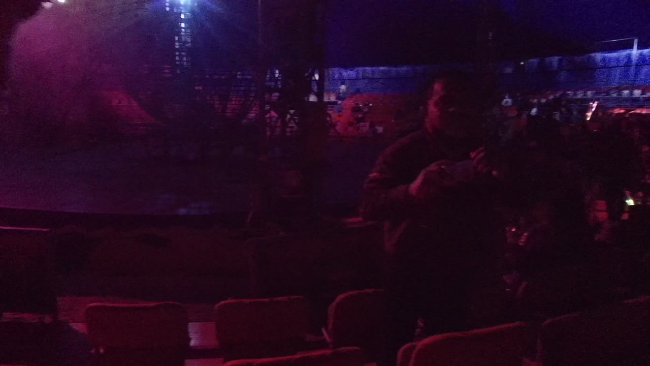 A dim San Jose del Cabo concert audience sits in red seats under purple and blue lighting, with shadowed people visible across the frame.