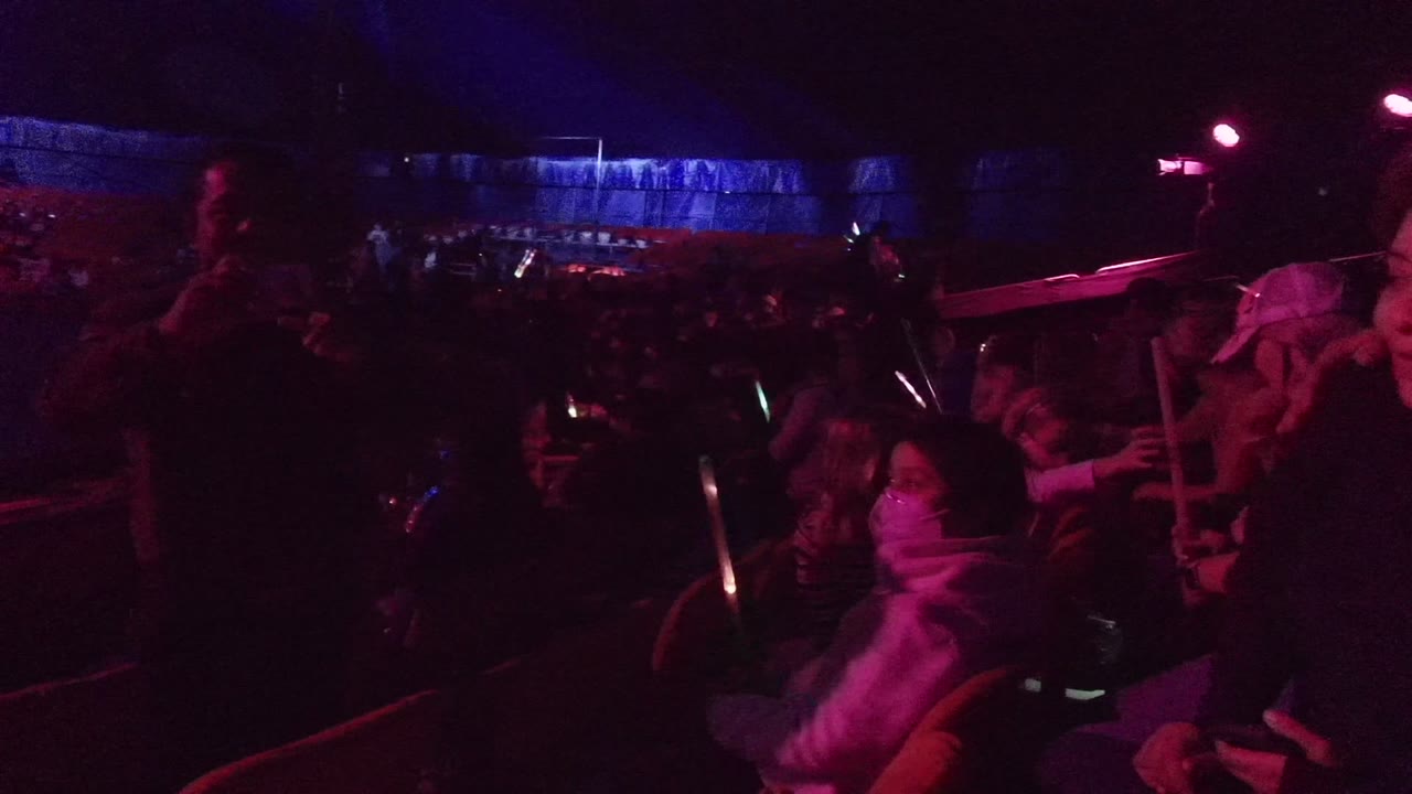 A closer view of the San Jose del Cabo audience shows people in red seats holding green glow sticks and phones under pink stage light.