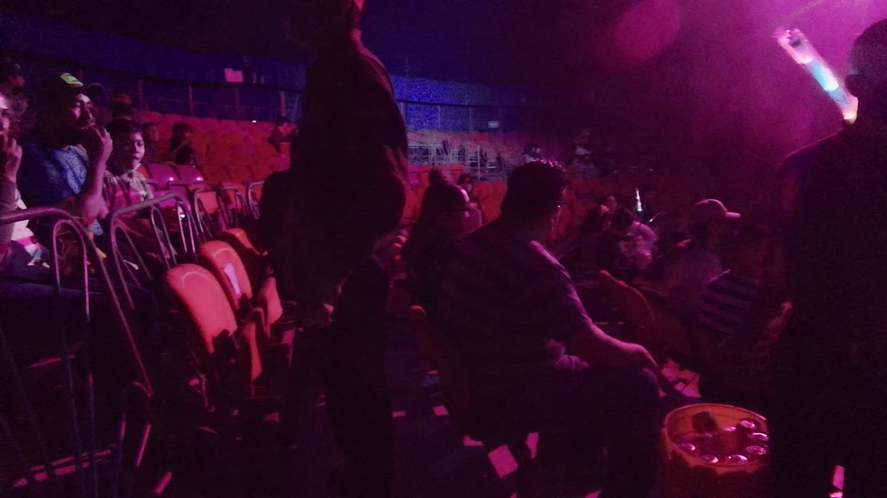 Audience seating in San Jose Del Cabo is shown in dim purple and pink light, with people seated and standing near rows of red chairs.