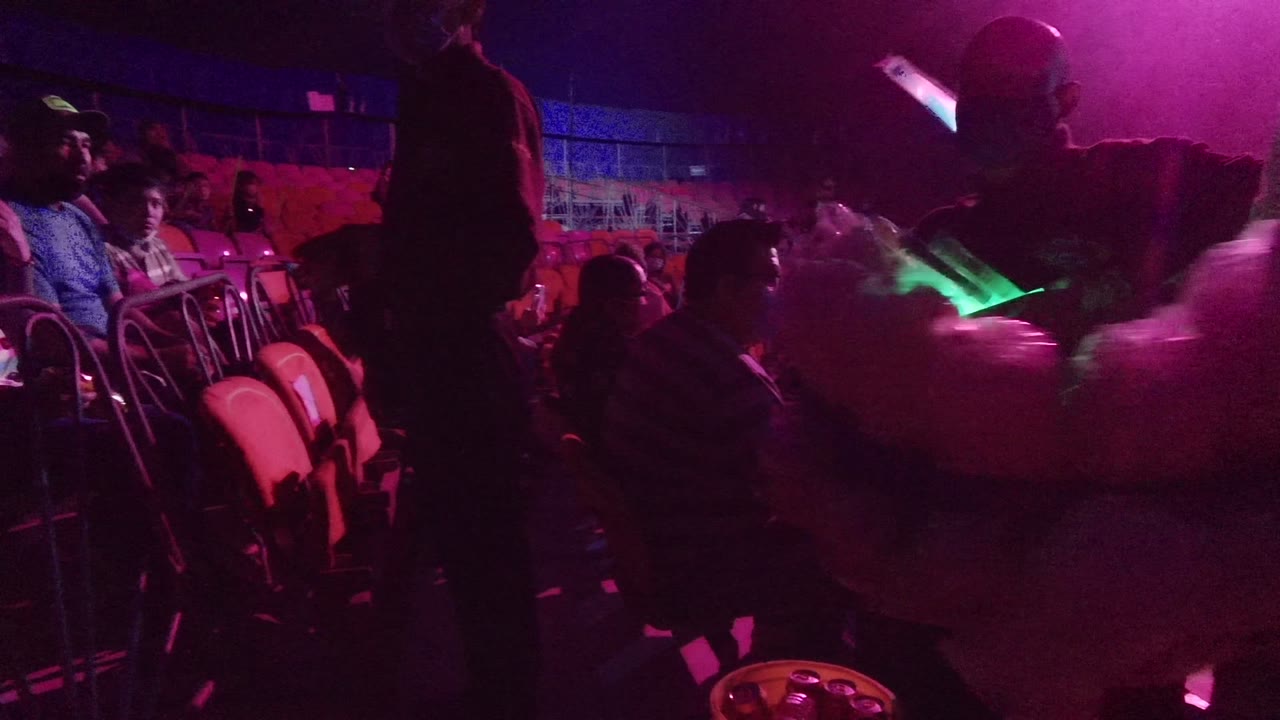 A silhouetted person stands close to the camera in San Jose Del Cabo while seated audience members and a green glowing object appear under pink-purple club light.