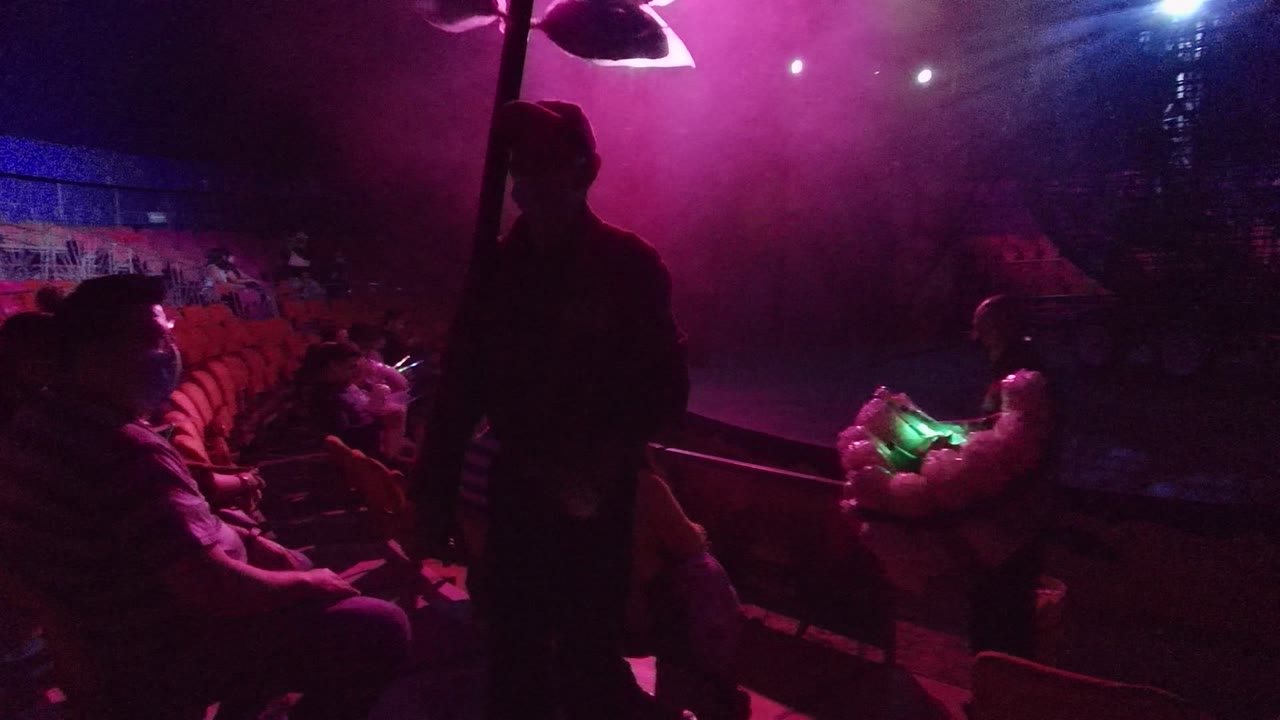 Audience members sit in red seats while a dark figure stands near the aisle in pink haze.