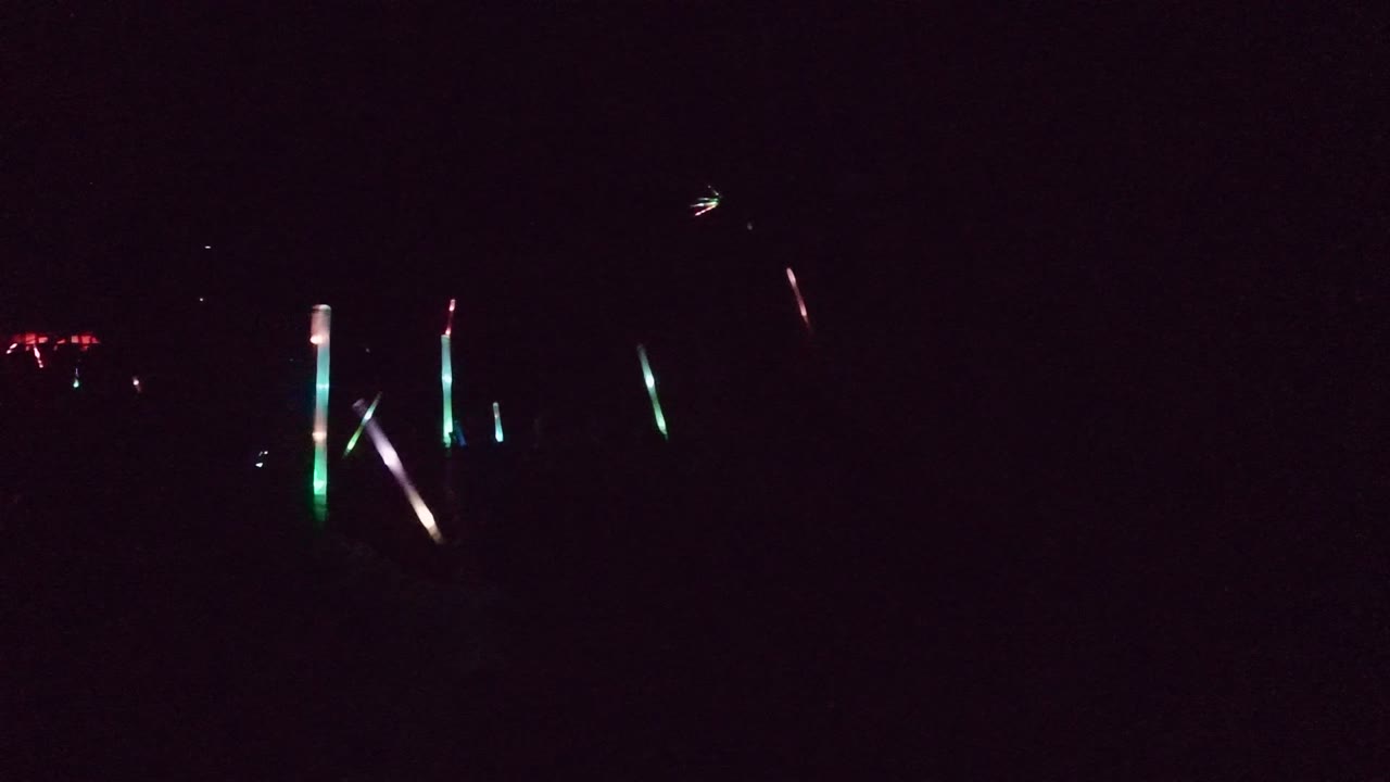 A very dark San Jose del Cabo event crowd with scattered green, pink, and white glow sticks visible against the black background.