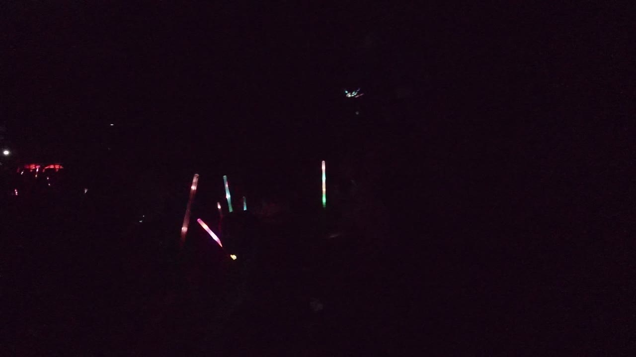 A mostly black San Jose del Cabo event frame with isolated colored glow sticks and faint distant red lights above the crowd.