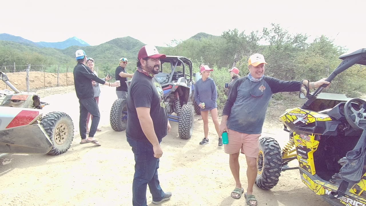 People in hats and casual clothing stand beside off-road vehicles on a sandy path in Los Cabos, with low desert hills and brush behind them.