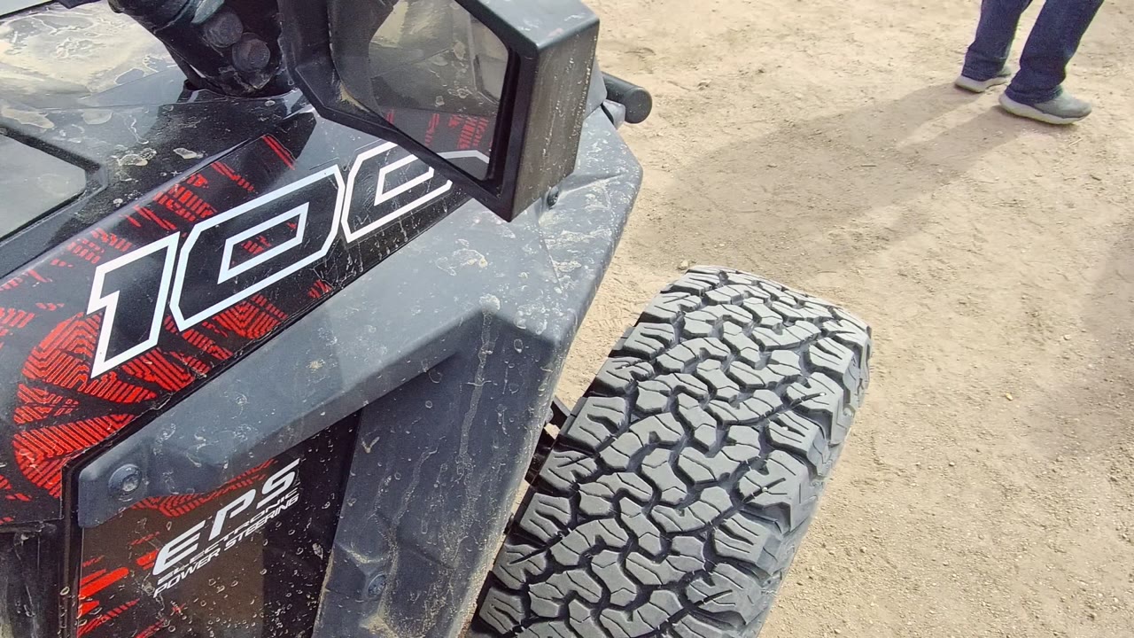 A low close view shows the off-road vehicle's side panel, handlebar, seat edge, cup holder, water bottle, and front tire, with parts of riders or passengers visible.