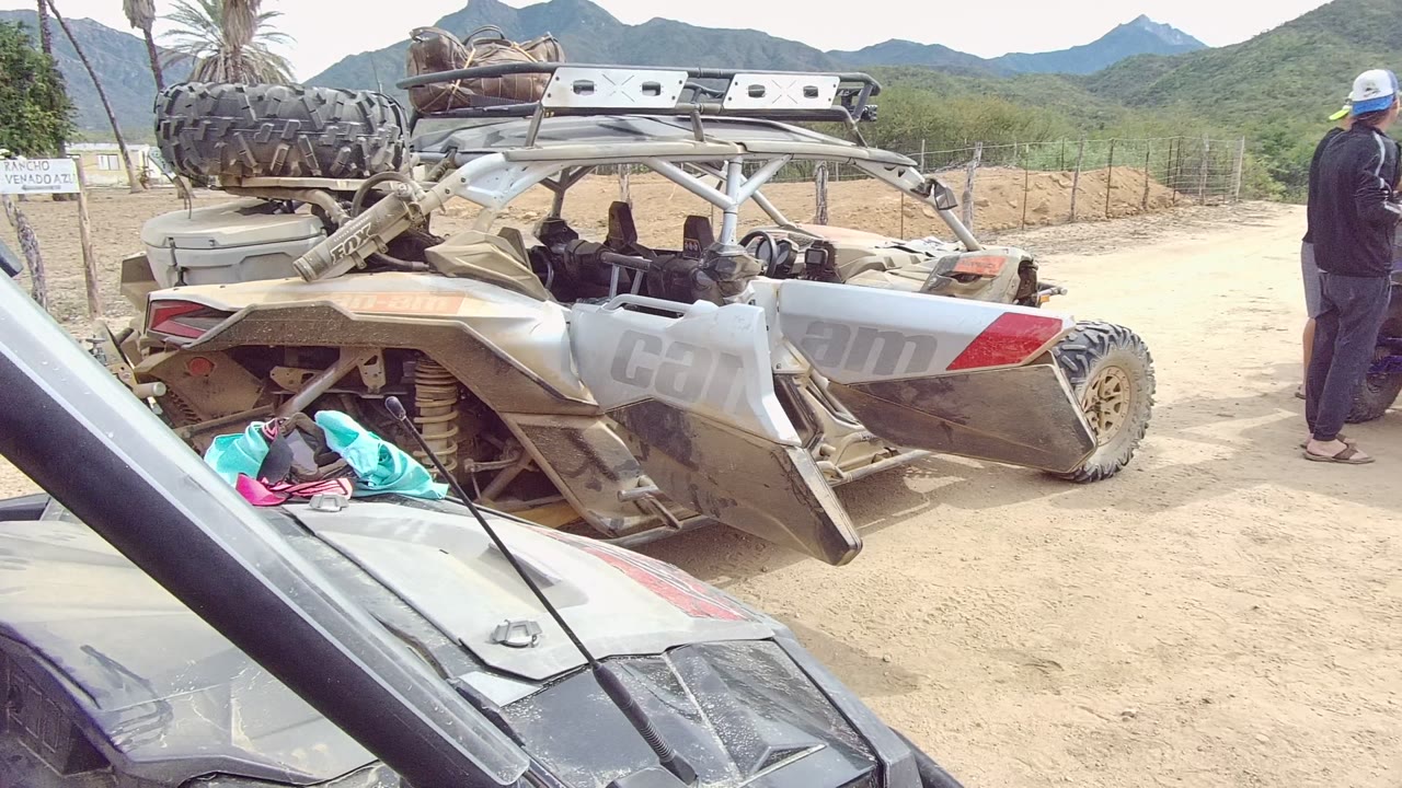 Wide Los Cabos frame showing a gray off-road vehicle lying on its side beside another parked vehicle, with sandy ground, scrub, mountains, and no clearly visible faces in the foreground.