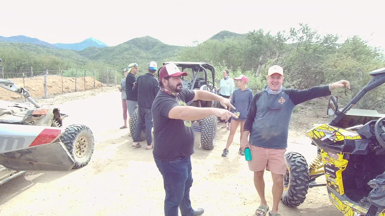 Two visible men in Los Cabos pose beside off-road vehicles on a sandy track, with one leaning toward the camera and flashing a peace sign while others stand behind them.