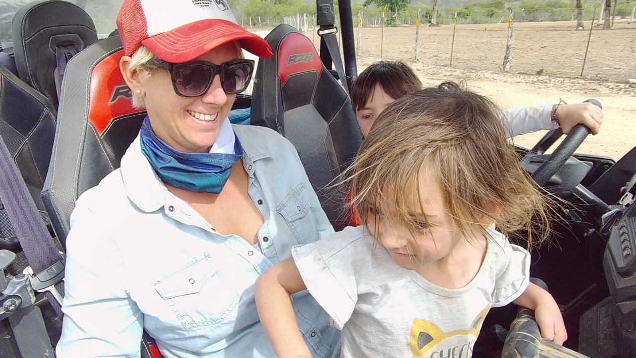 Woman in sunglasses and a red-and-white cap smiles in an off-road vehicle with a young child in front and a boy partly visible at the wheel in Los Cabos, Mexico.