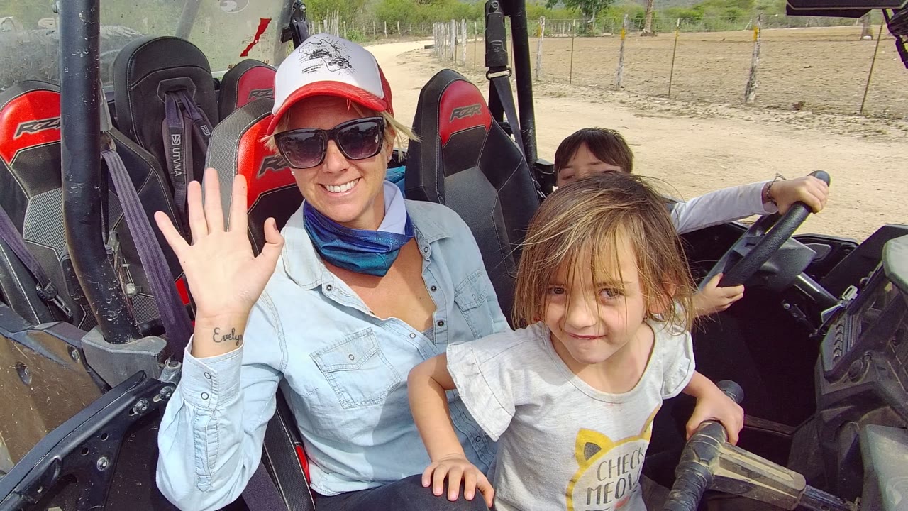 Woman in sunglasses raises one hand in a wave beside a young child, with the boy still at the steering wheel and dry Los Cabos terrain behind the vehicle.