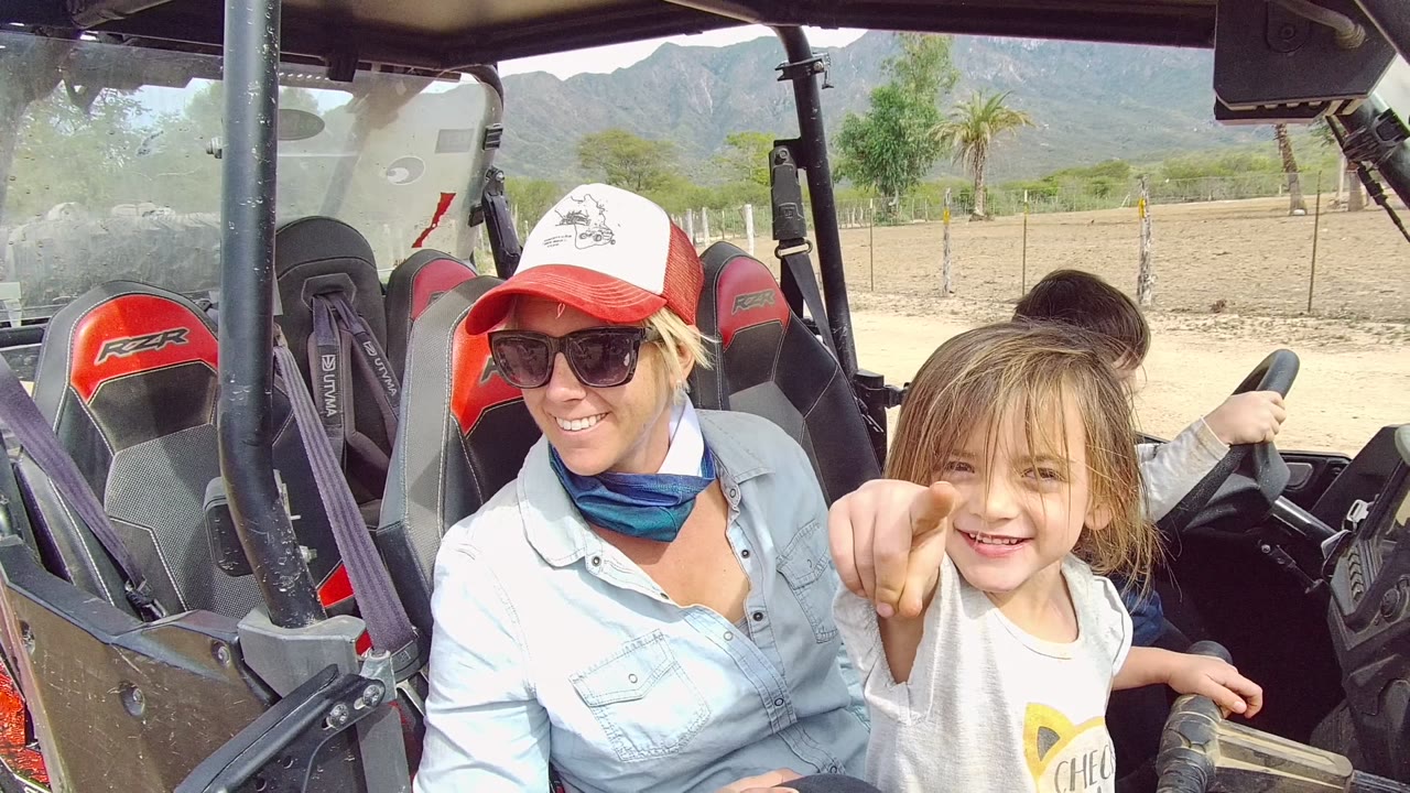 At 0:00 in Los Cabos, Baja California Sur, a woman in a red cap and sunglasses sits beside a smiling child in an off-road buggy, with another child at the wheel behind them.