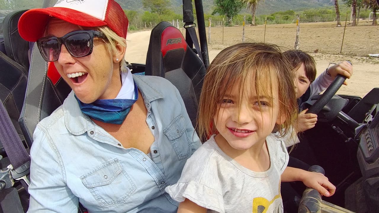 At 0:02 in Los Cabos, Baja California Sur, the woman reacts with her mouth open while the child looks toward camera and another child holds the steering wheel in back.
