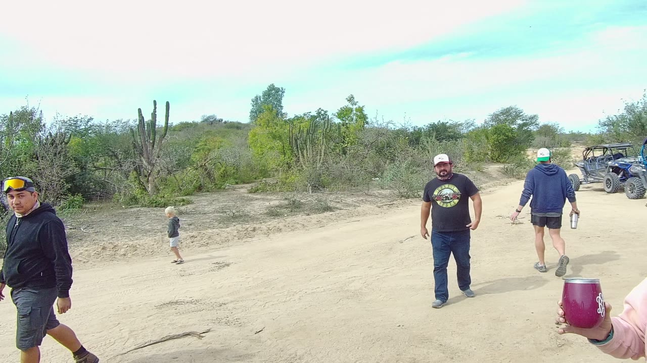 A man in a dark hoodie walks near a child on a pale sandy road in Los Cabos, with desert scrub, tall cacti, and low hills across the background.