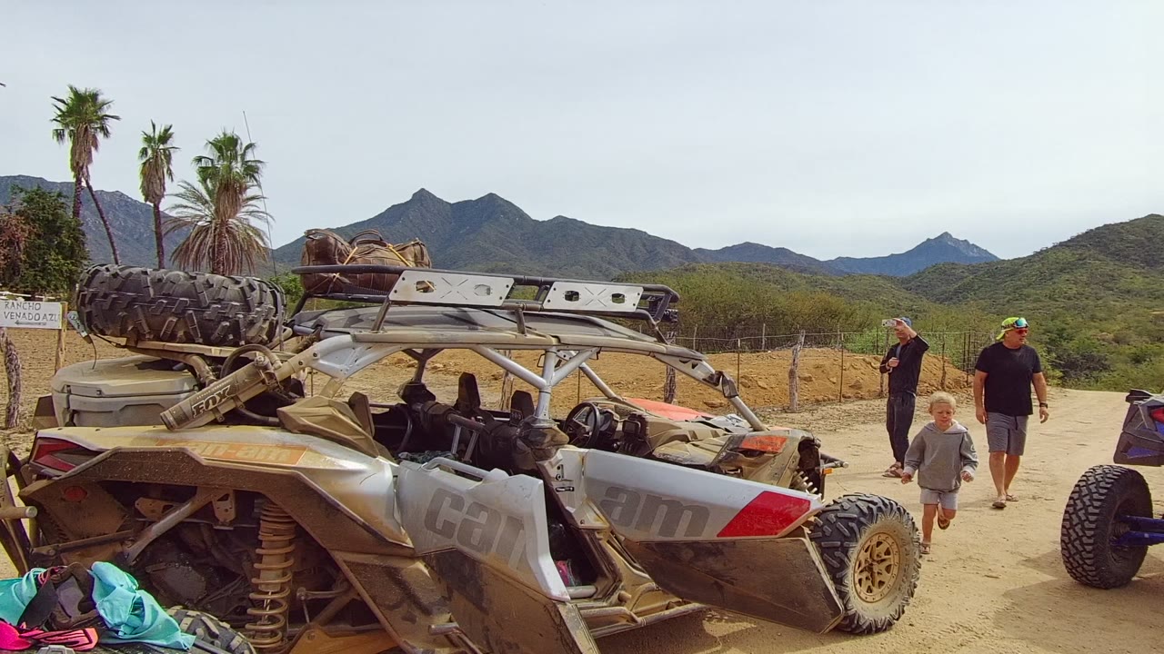 A silver off-road vehicle fills the foreground in Los Cabos, Baja California Sur, with adults and a child nearby, palm trees, scrub, and mountains behind it.
