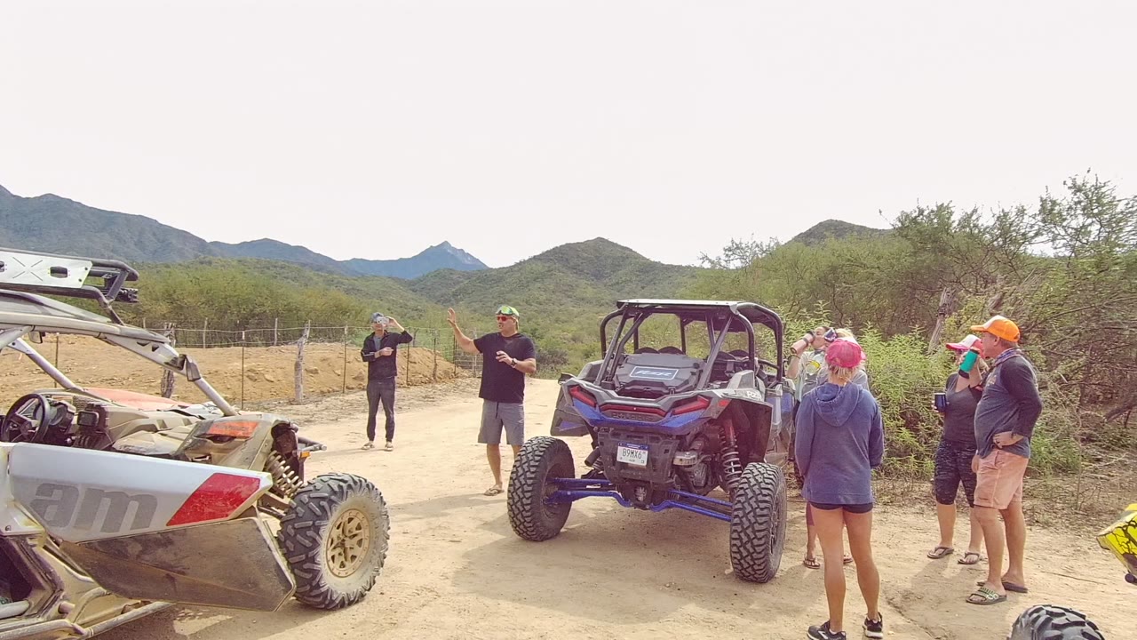 A blue off-road vehicle sits near the center of the Los Cabos desert road as a child moves across the foreground and adults stand around the vehicles.