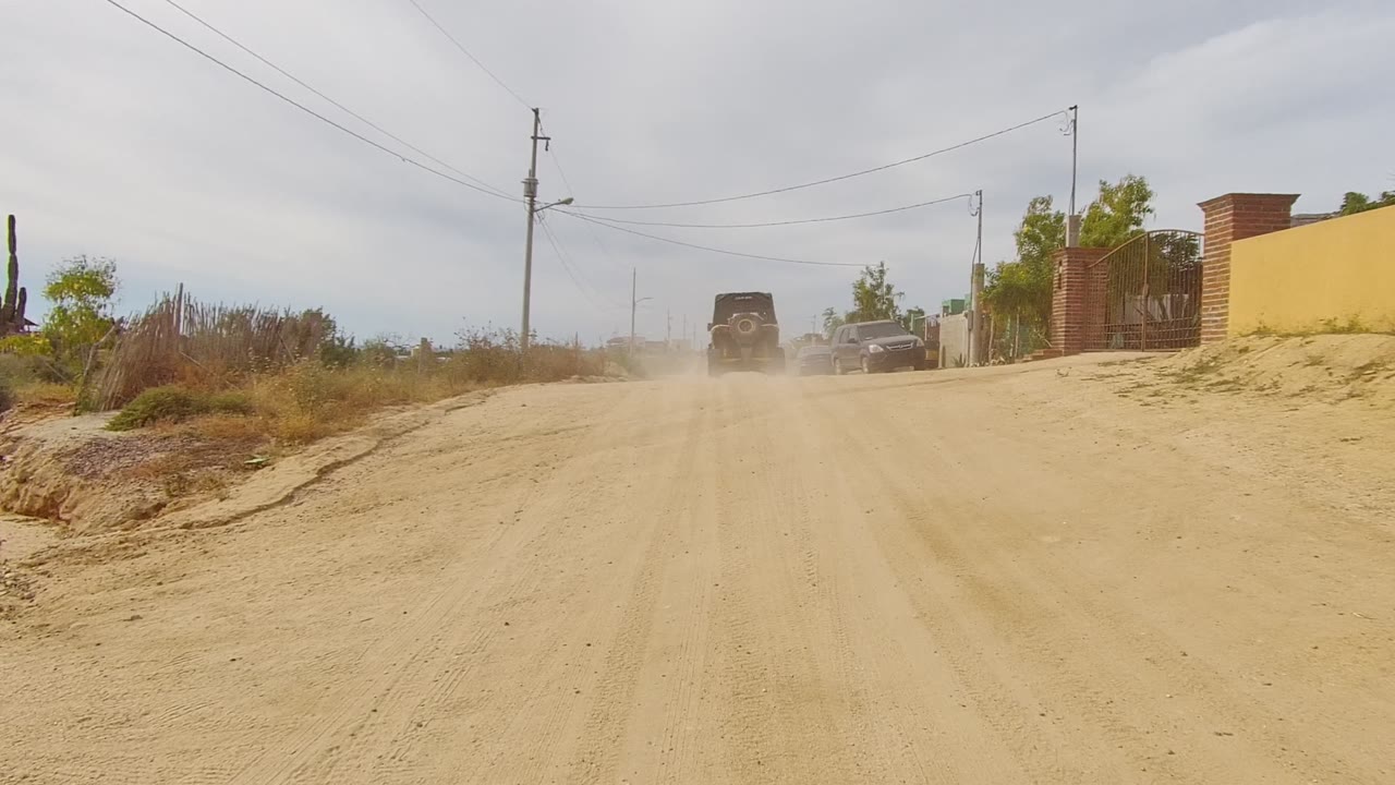 A vehicle drives away on a sandy unpaved road in Los Cabos, Baja California Sur, with dry brush, utility poles, parked cars, low buildings, and no visible people.