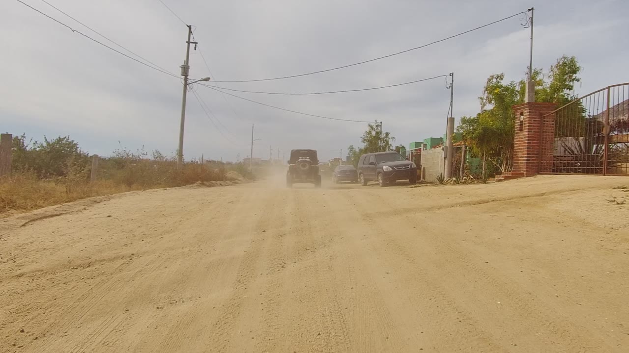 Dusty road with a vehicle ahead and parked cars near roadside buildings.