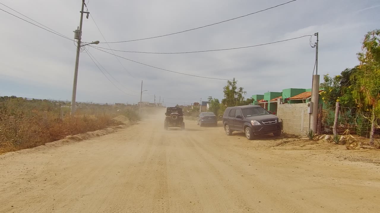A vehicle recedes through dust on an unpaved Los Cabos road, with a parked dark SUV, green roadside buildings, dry brush, power lines, and no visible people.