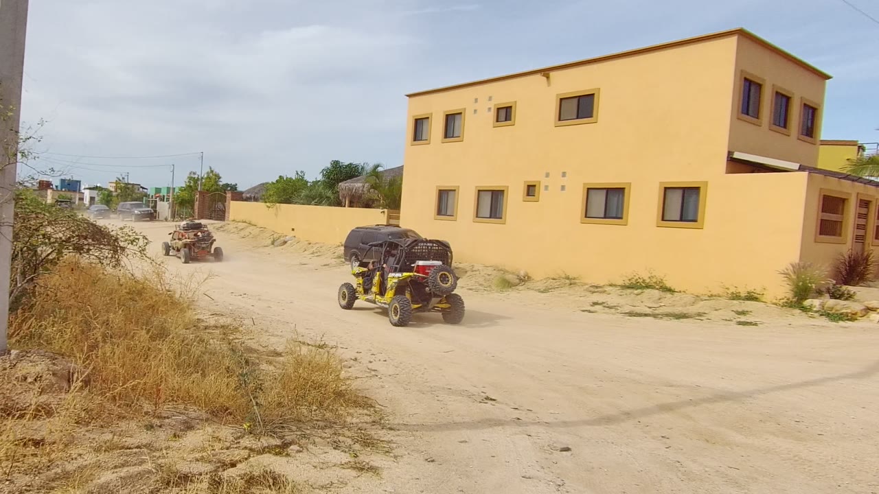 Yellow off-road vehicle drives on a sandy road in Los Cabos beside dry brush, a yellow building, and a parked black SUV, with no visible people.