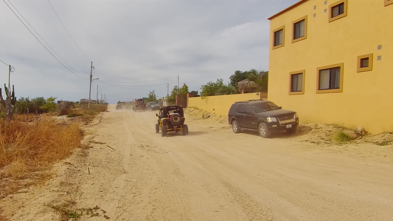 The off-road vehicle continues away on the dusty Los Cabos dirt road, leaving the black SUV and yellow building behind in the right foreground, with no visible people.