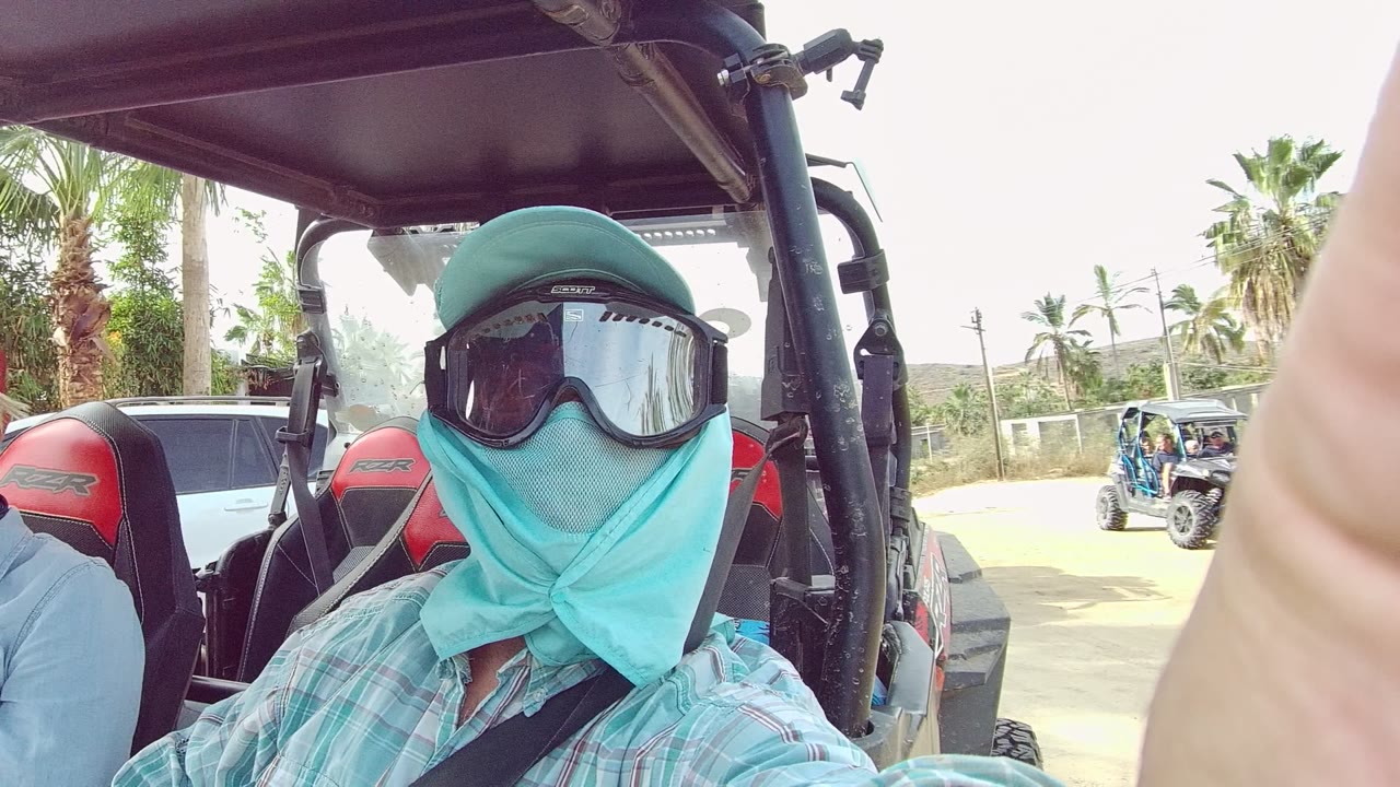 Close view of a masked and goggled rider inside an open UTV in Los Cabos, with a dusty roadside and parked vehicle partly visible behind them.