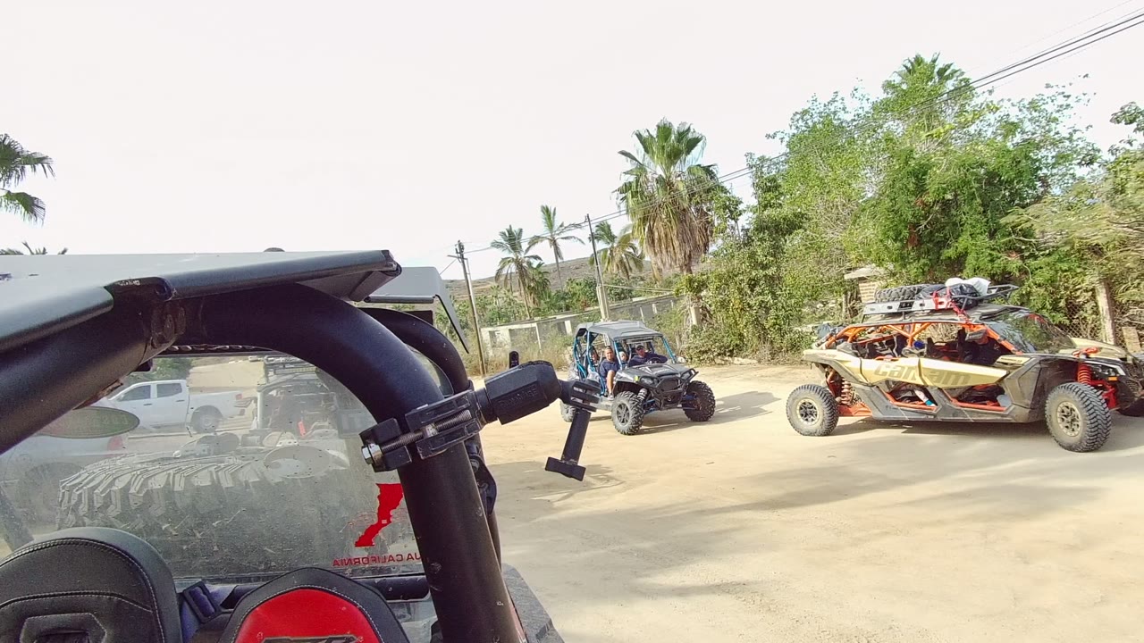 Two dune buggies travel along a dusty road in Los Cabos, with the filming vehicle's roll bar and seat edge in the foreground and palm trees behind.
