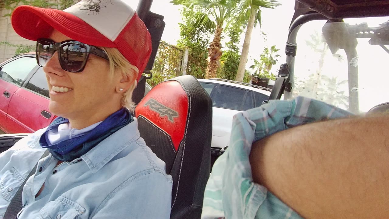 A woman wearing sunglasses, a red cap, a neck gaiter, and a light denim shirt sits smiling inside an off-road vehicle in Los Cabos.