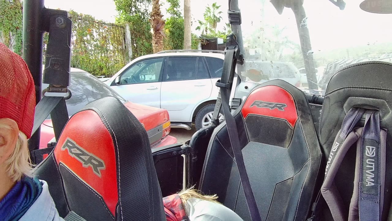 Inside a RZR UTV in Los Cabos, the first frame shows red and black seats, harness straps, a partial person in a red cap at left, and parked cars behind the vehicle.