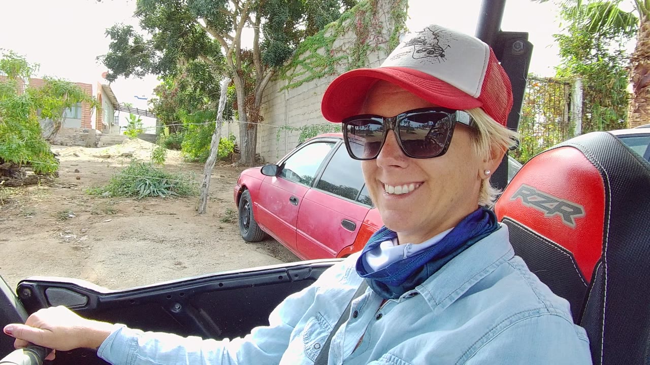 At 0:00, a person in a red and white baseball cap and sunglasses drives an open vehicle on a residential dirt road in Los Cabos, with a red car and trees behind them.