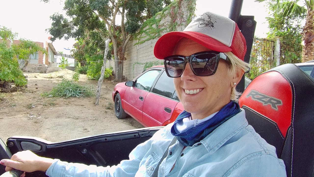 Smiling driver in sunglasses seated in an open vehicle beside a red car.