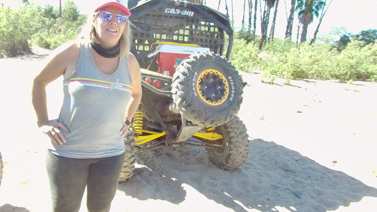 Woman facing camera near a Can-Am vehicle on a sunny sandy path.
