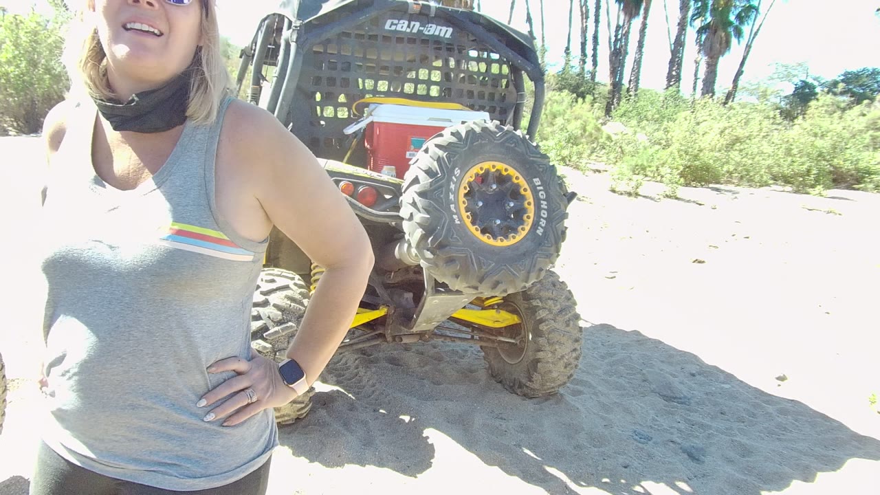 In Los Cabos, Baja California Sur, a woman in sunglasses and a red cap stands partly cropped beside a Can-Am ATV on bright sandy ground.